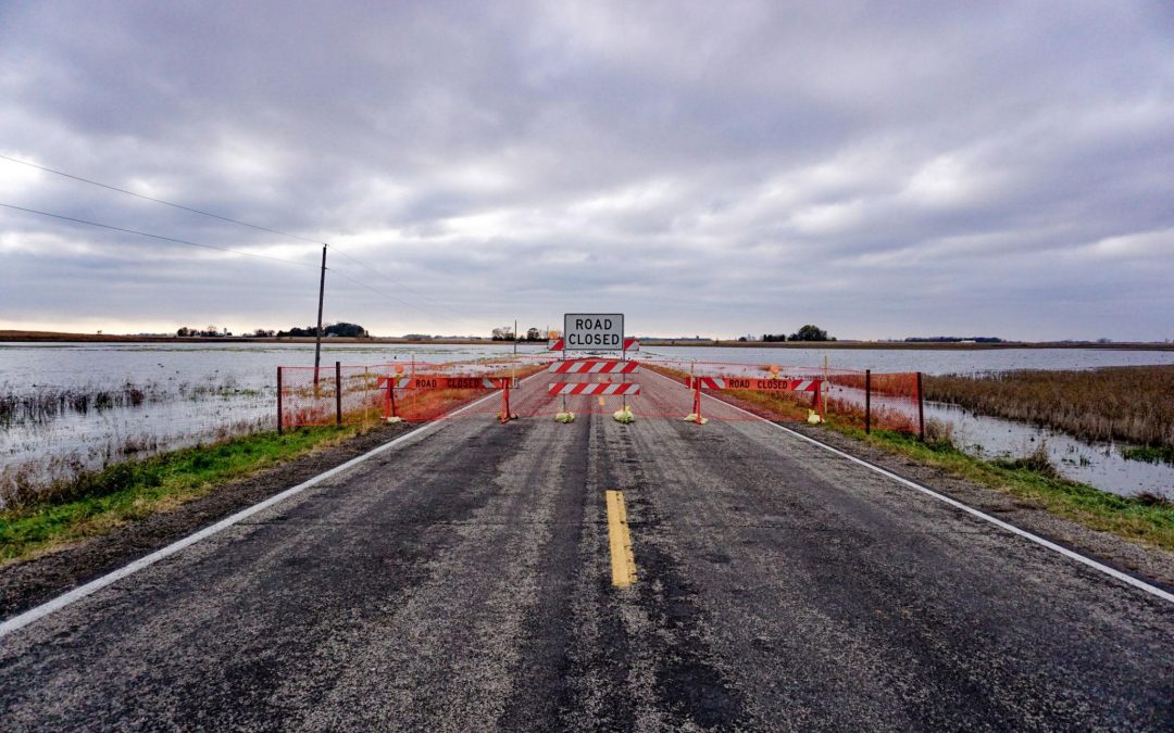 Floodwater covering a street with road closure barriers and trees nearby. Nebraska Seeks FEMA Aid After $23M in Flood Damage.
